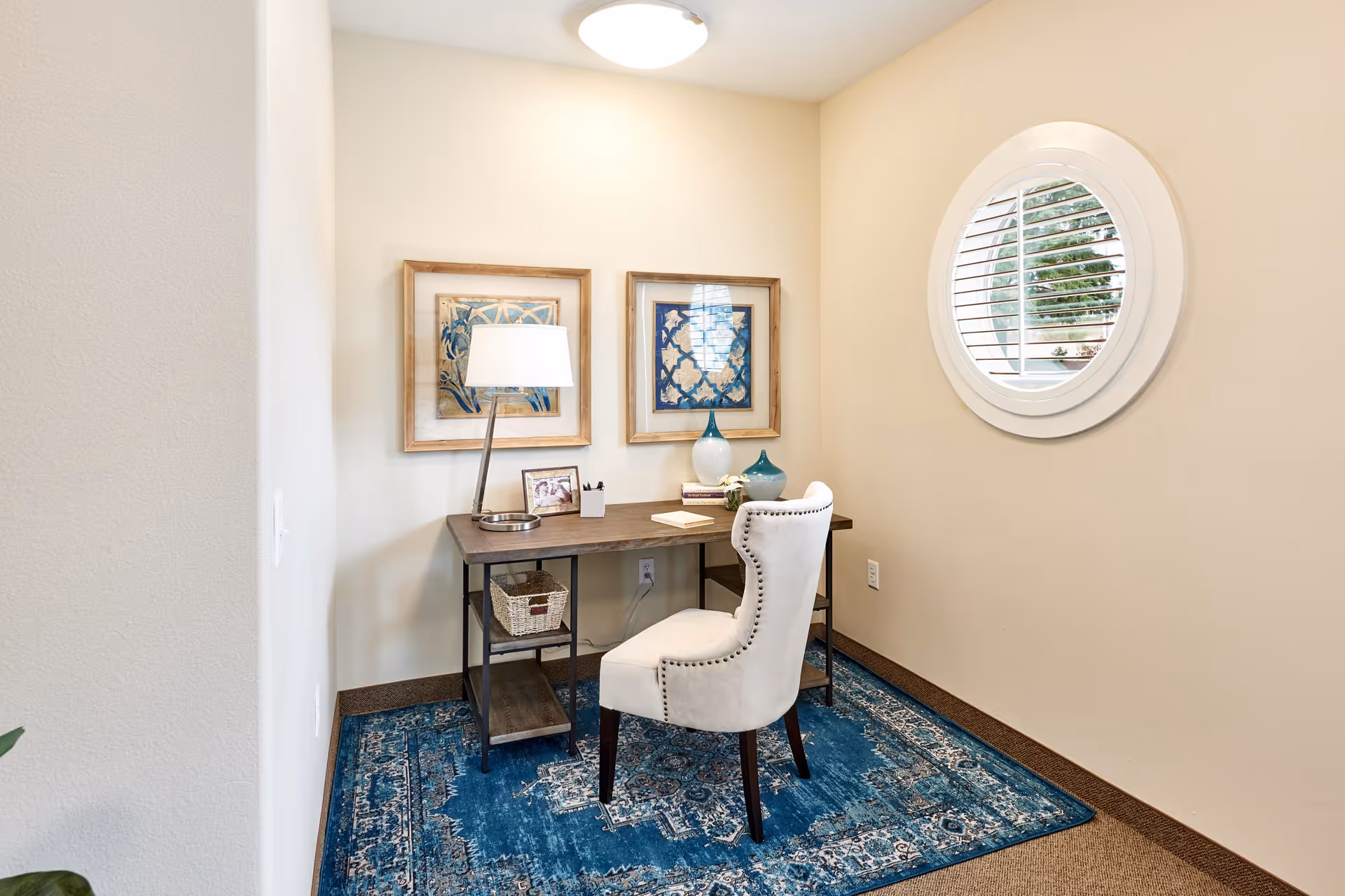 A small office nook with a wooden desk and a white upholstered chair on a blue patterned rug. The desk has a lamp, framed photos, books, and decorative vases. Two framed artworks hang on the wall above the desk, and a round window with white shutters is on the adjacent wall.