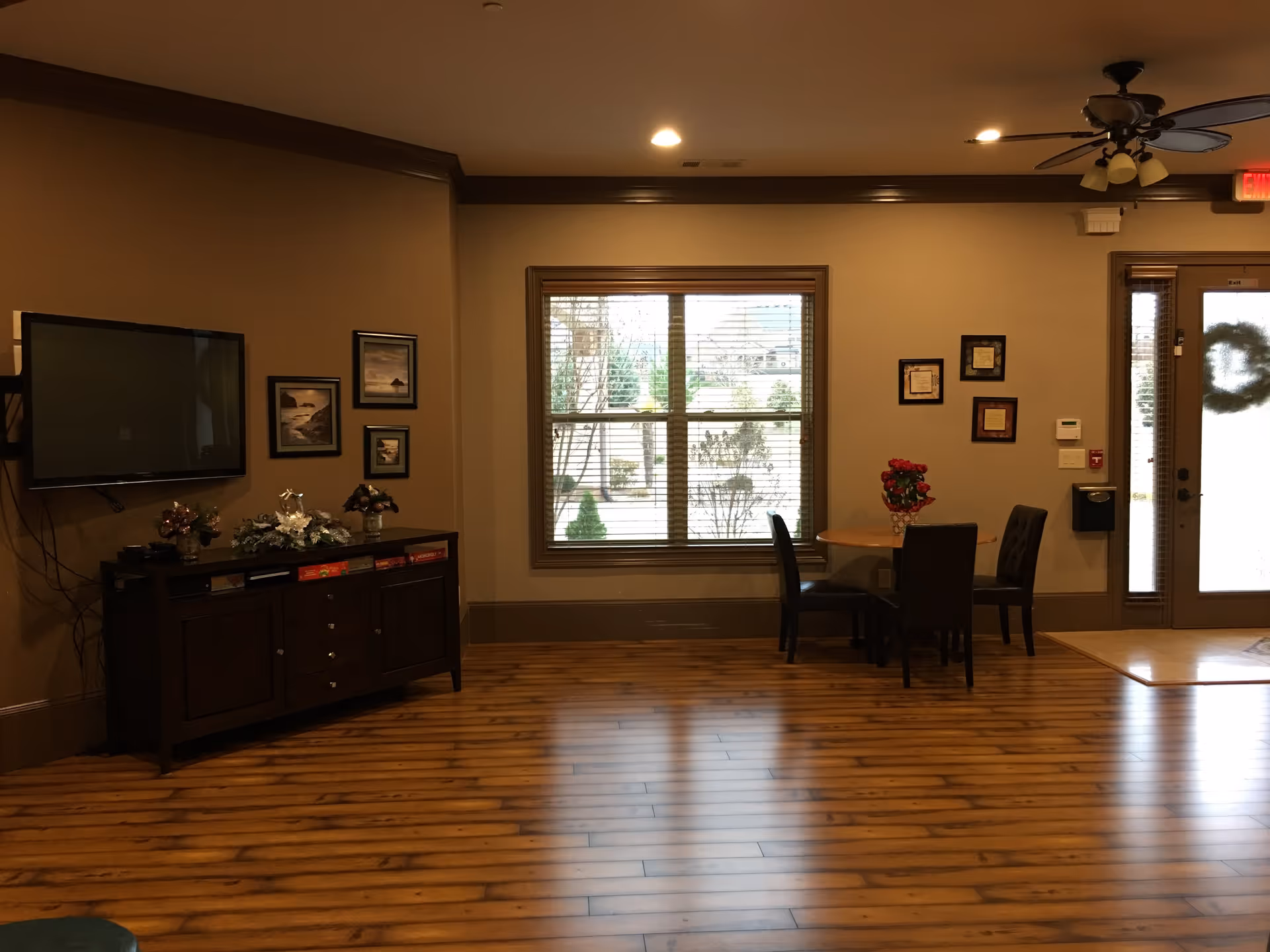 Interior view of a senior living community common area with wooden flooring, a wall-mounted TV above a dark wooden cabinet, framed pictures on the wall, a window with blinds showing outdoor greenery, a small round table with four chairs, and a door with a wreath.