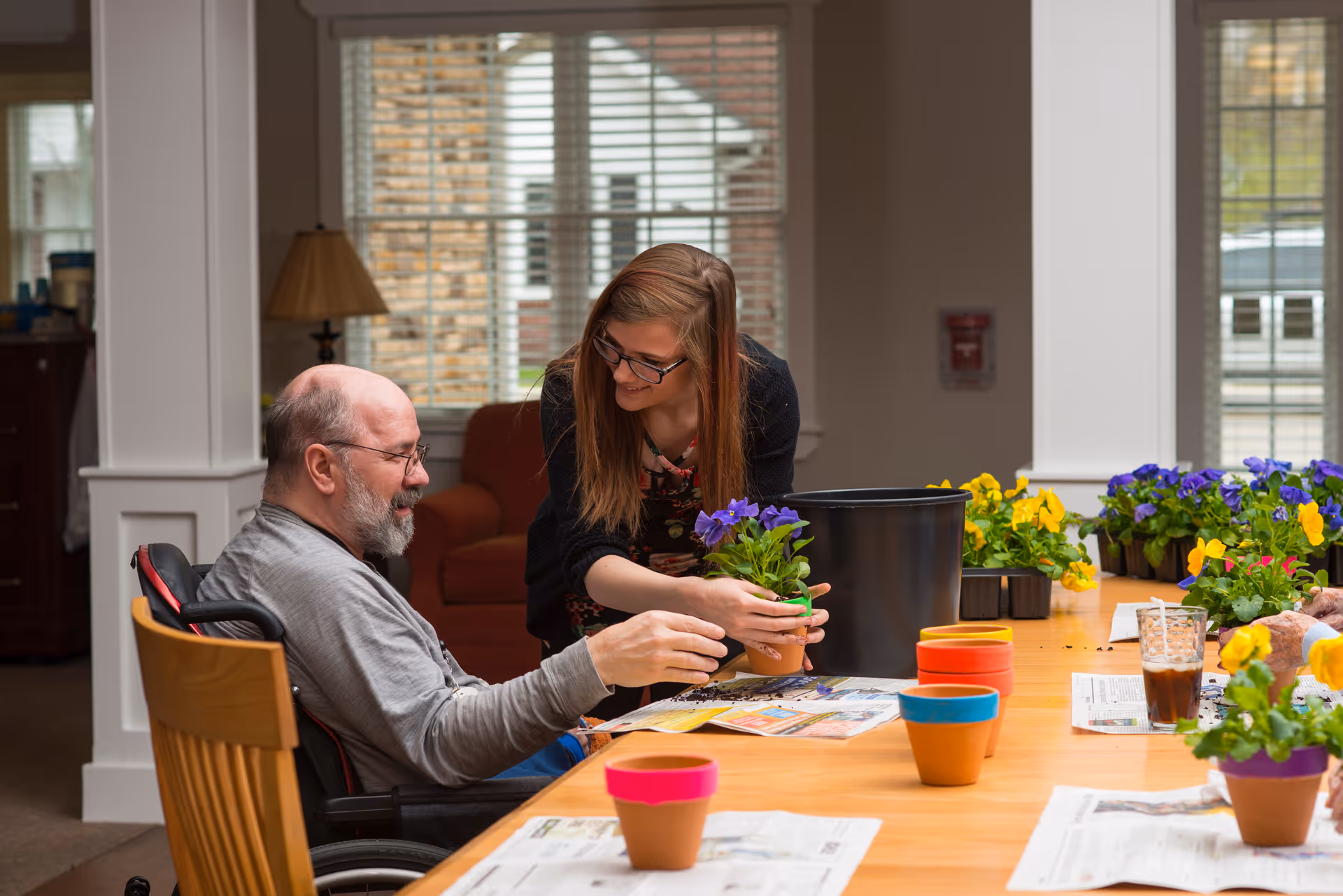An elderly man in a wheelchair and a younger woman with glasses are sitting at a table indoors, engaging in a gardening activity. The woman is handing a small potted plant with purple flowers to the man. The table is covered with newspapers and several colorful flower pots, with more potted flowers in the background. The room has large windows with blinds and a cozy atmosphere.