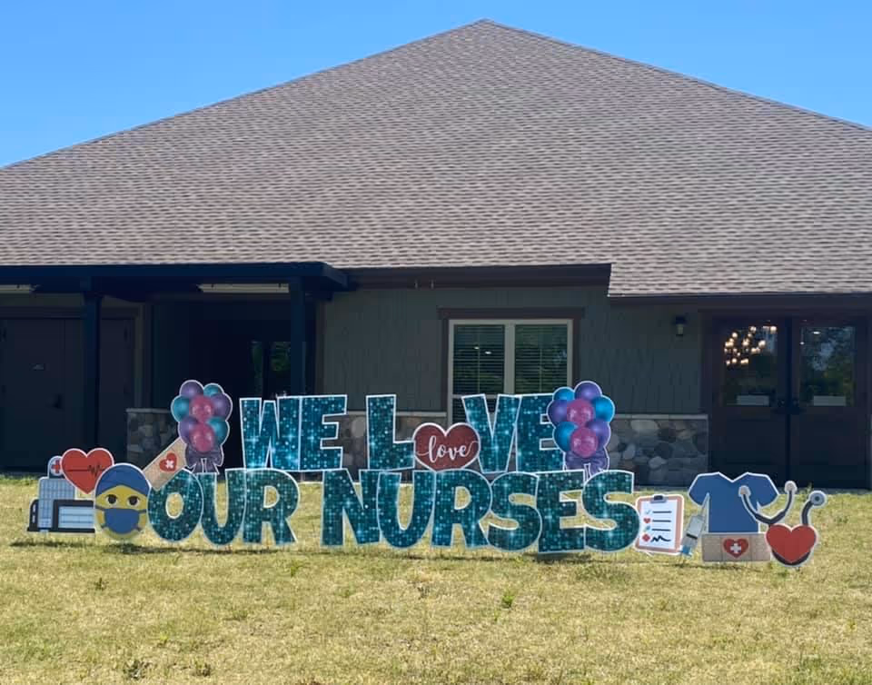 Outdoor view of a building with a large decorative sign on the grass in front that reads 'WE love OUR NURSES' with colorful balloons and healthcare-related icons such as a nurse emoji, medical clipboard, scrubs, and stethoscope.