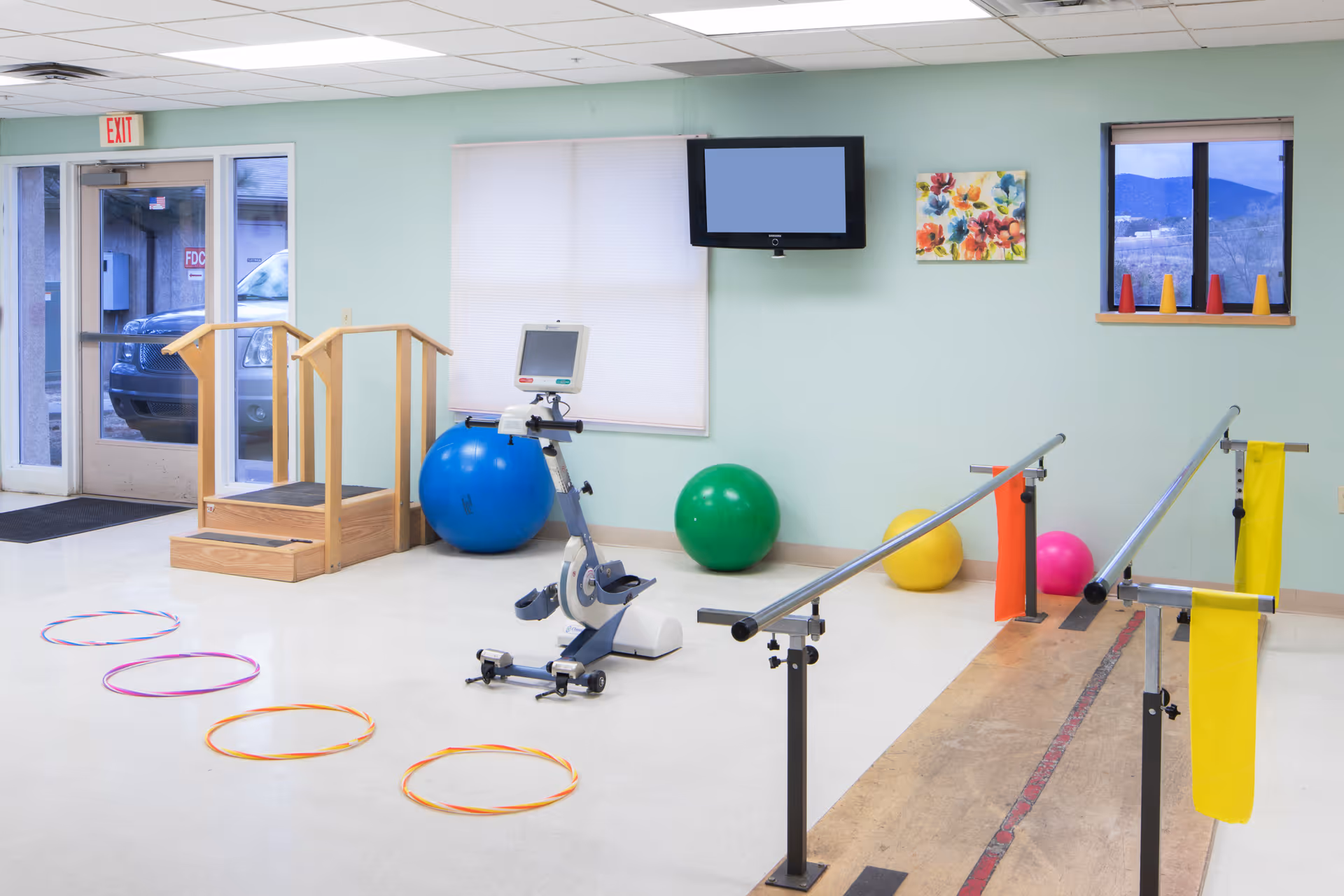 Bright therapy/exercise room with parallel bars, an exercise bike, large stability balls, a step platform, hula hoops, and a wall-mounted TV.