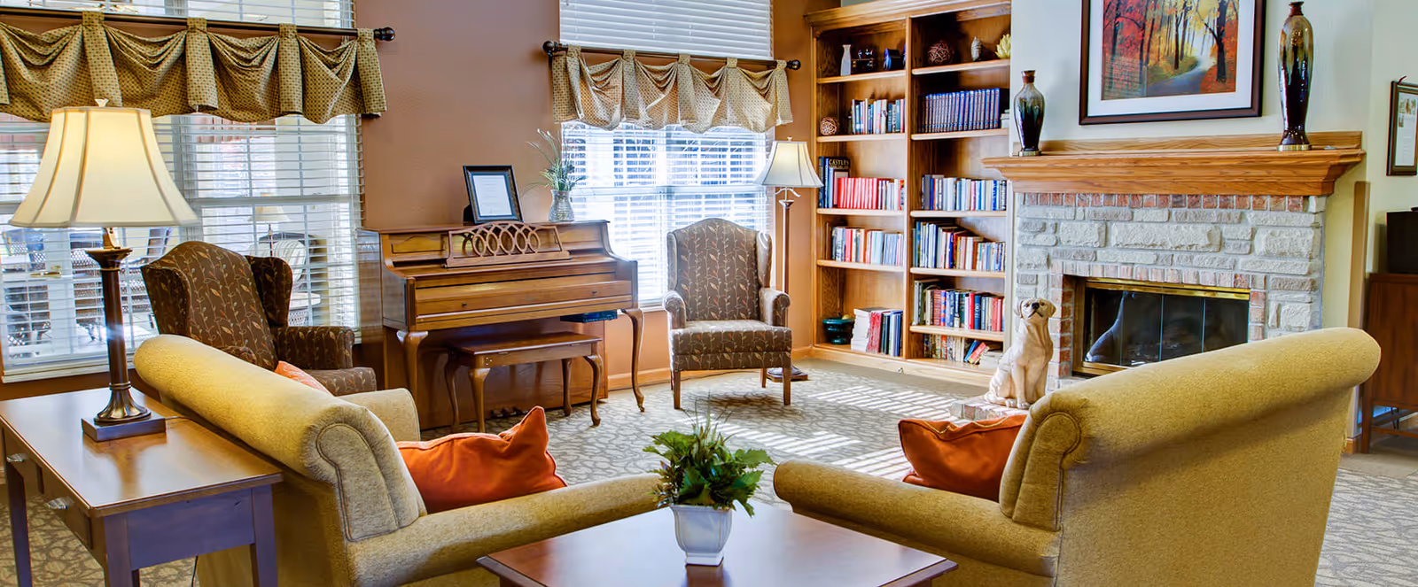 A cozy living room with two beige sofas facing each other, each with orange throw pillows. There are two patterned armchairs near a wooden piano by the window. A wooden bookshelf filled with books is next to a stone fireplace with a framed painting above it. A small table with a plant is in the center, and the room is well-lit by natural light coming through the windows with valance curtains.