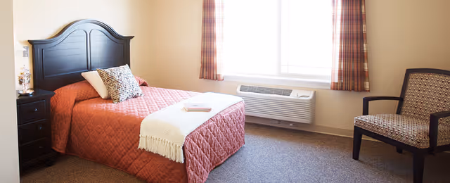 A bright bedroom with a single bed covered in a red quilt and a white throw blanket. The bed has a dark wooden headboard and a decorative pillow. Next to the bed is a black nightstand with a small lamp and decorative item. A window with plaid curtains lets in natural light. There is a patterned armchair to the right of the window and a wall-mounted air conditioning unit below the window.