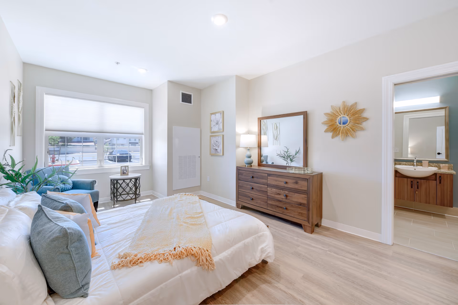 A well-lit bedroom featuring a comfortable bed with decorative pillows, a wooden dresser, and a mirror. A cozy chair and a small table are positioned near a large window, allowing natural light to fill the space. The room has a warm color palette and modern decor.