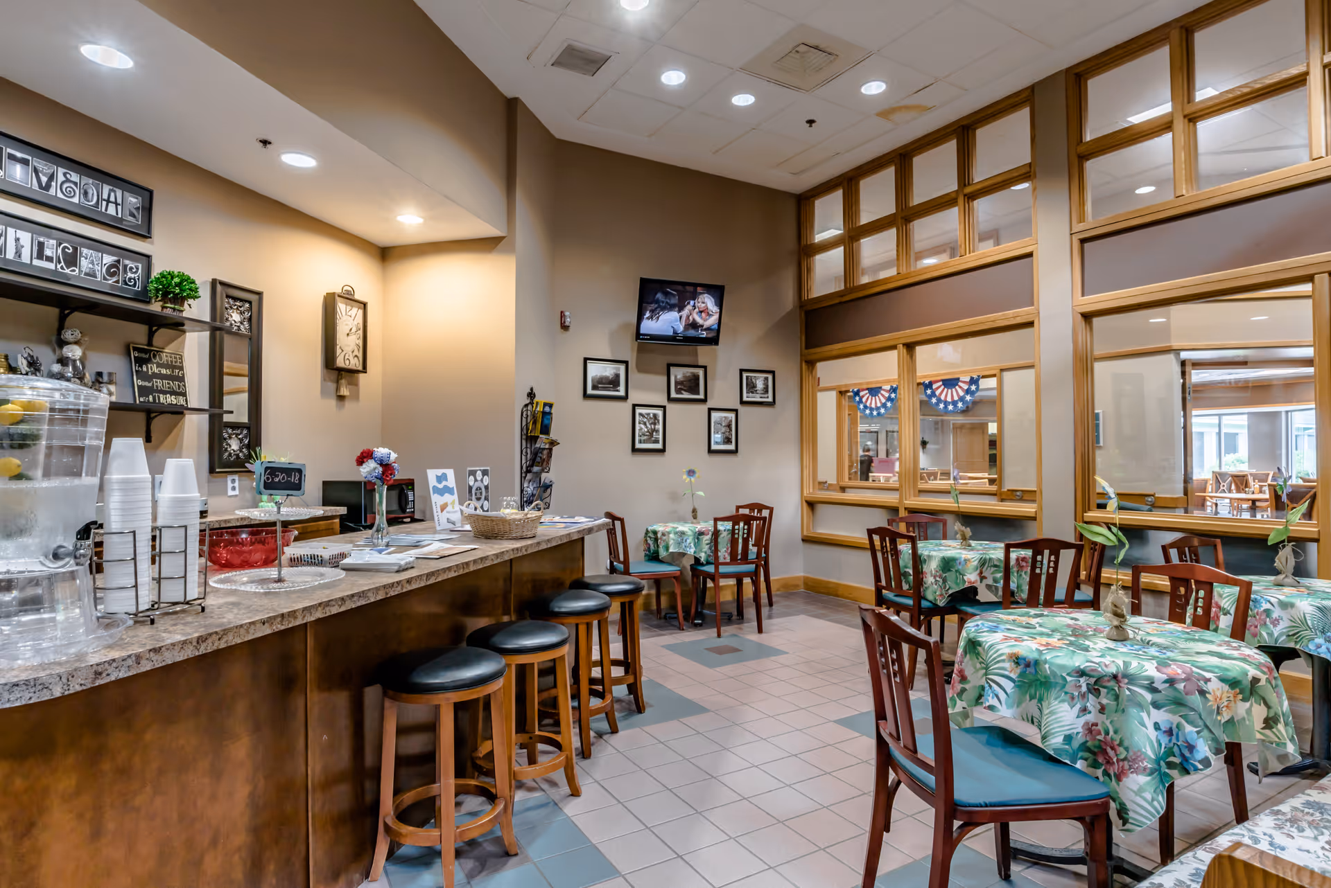 Interior view of a dining area in a senior living facility with several tables covered in floral tablecloths and wooden chairs. A counter with stools is on the left side, featuring a water dispenser, cups, and decorative items. The walls are adorned with framed pictures and a clock, and a television is mounted on the far wall. Large windows with wooden frames separate this room from another area.
