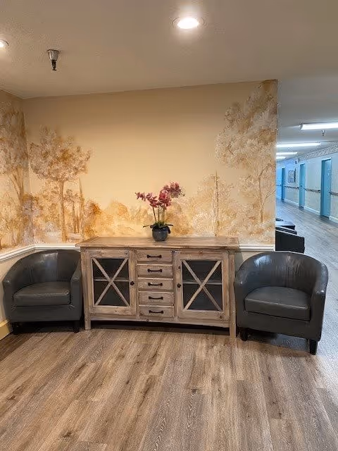A cozy corner in a senior living facility with two dark gray armchairs flanking a wooden cabinet with drawers and glass-paneled doors. A potted plant with pink flowers sits on top of the cabinet. The walls have a soft mural of trees in warm tones, and a hallway with light wood flooring extends to the right.