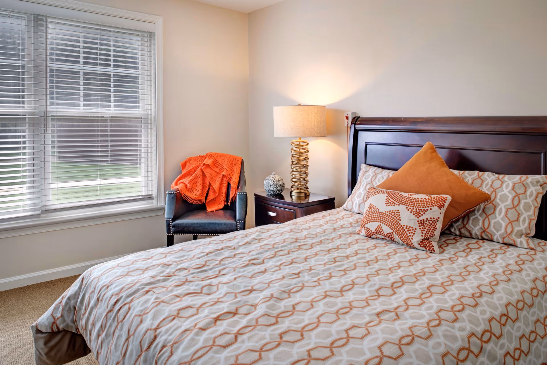 Bedroom with a patterned orange-and-white bed, wooden headboard, nightstand with lamp, and a chair by a large window.