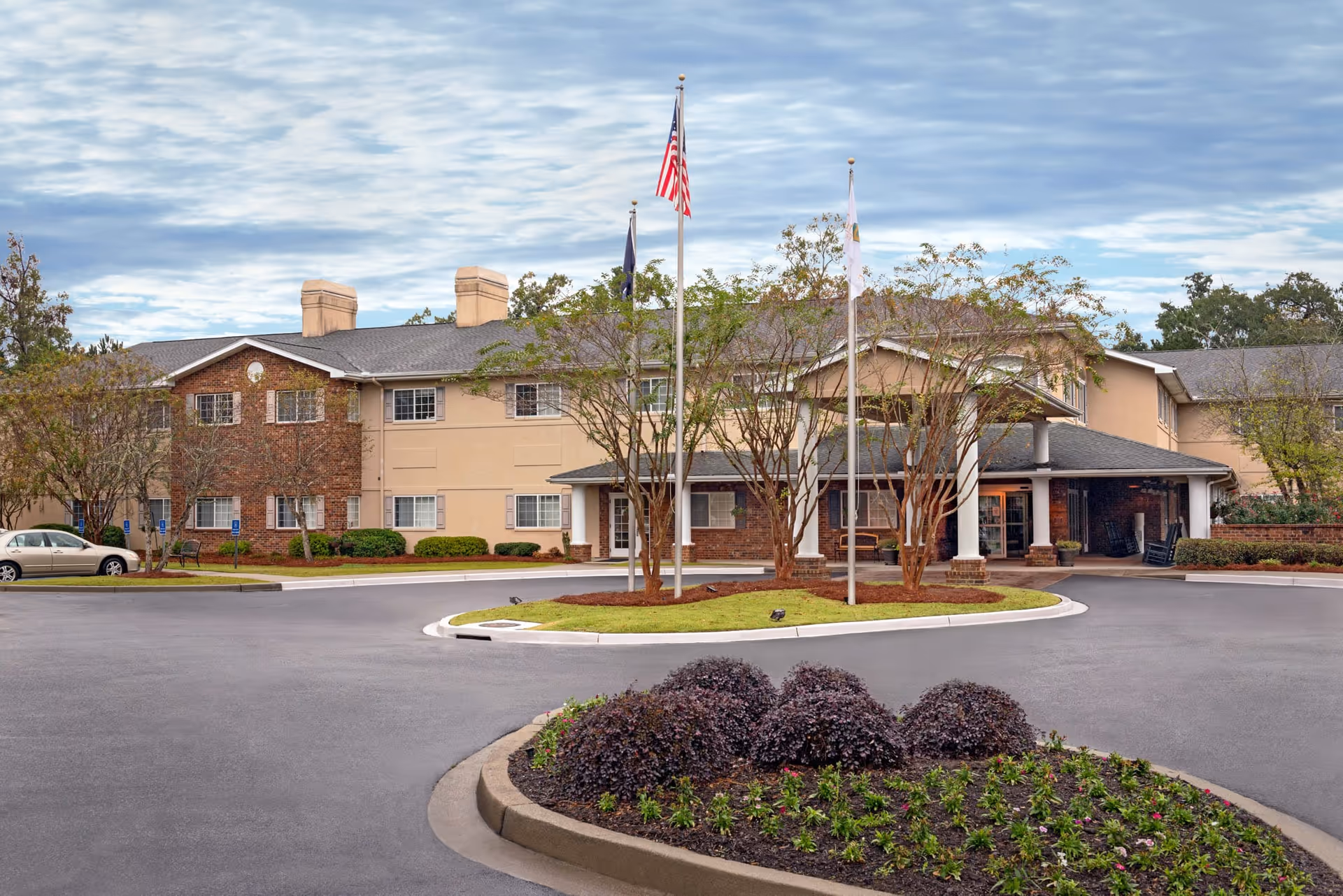 Exterior view of The Bridge at Charleston senior living facility showing a two-story building with a covered entrance, three flagpoles with flags, landscaped bushes and trees, and a circular driveway.