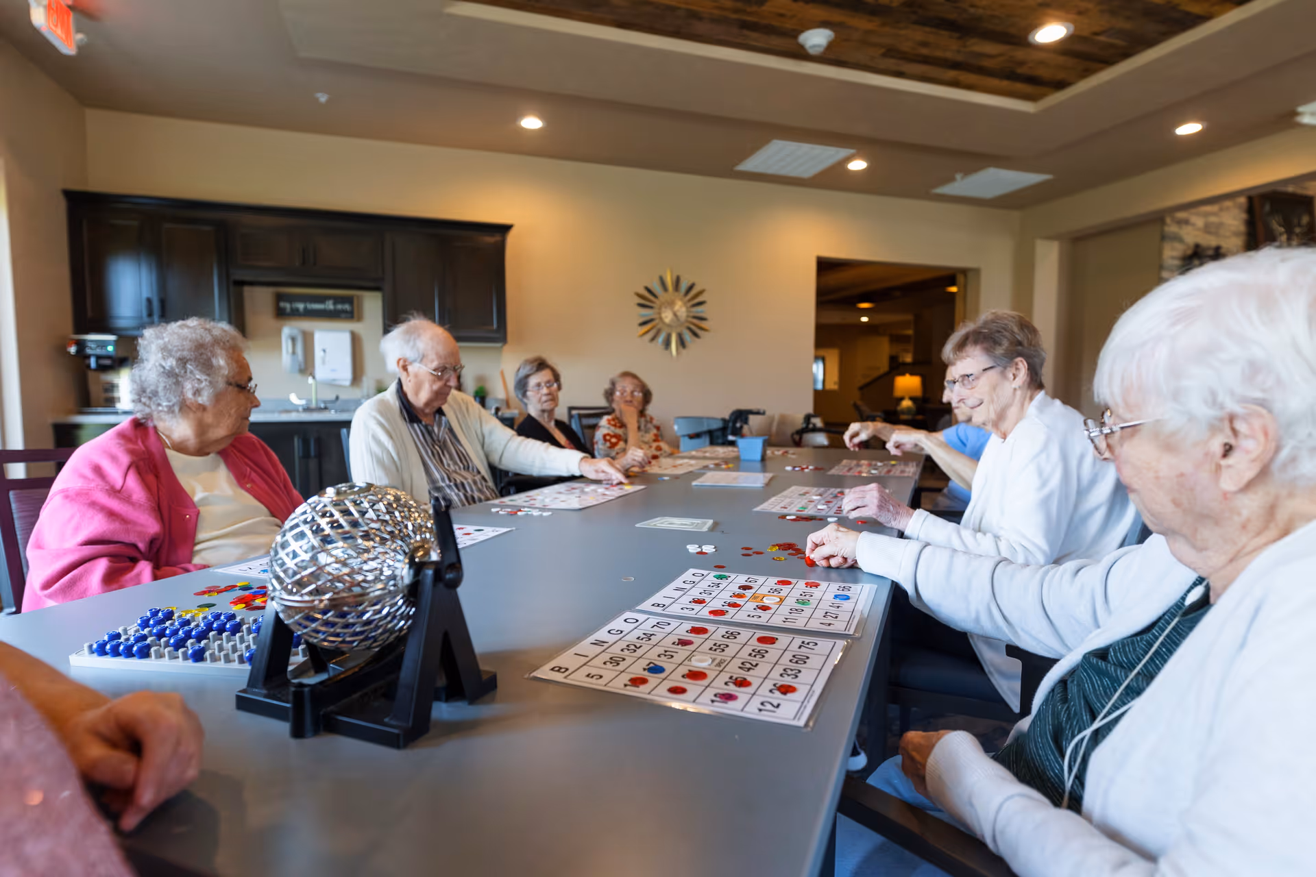 A group of elderly people sitting around a large table playing bingo in a well-lit room with wooden ceiling accents and a decorative wall clock.