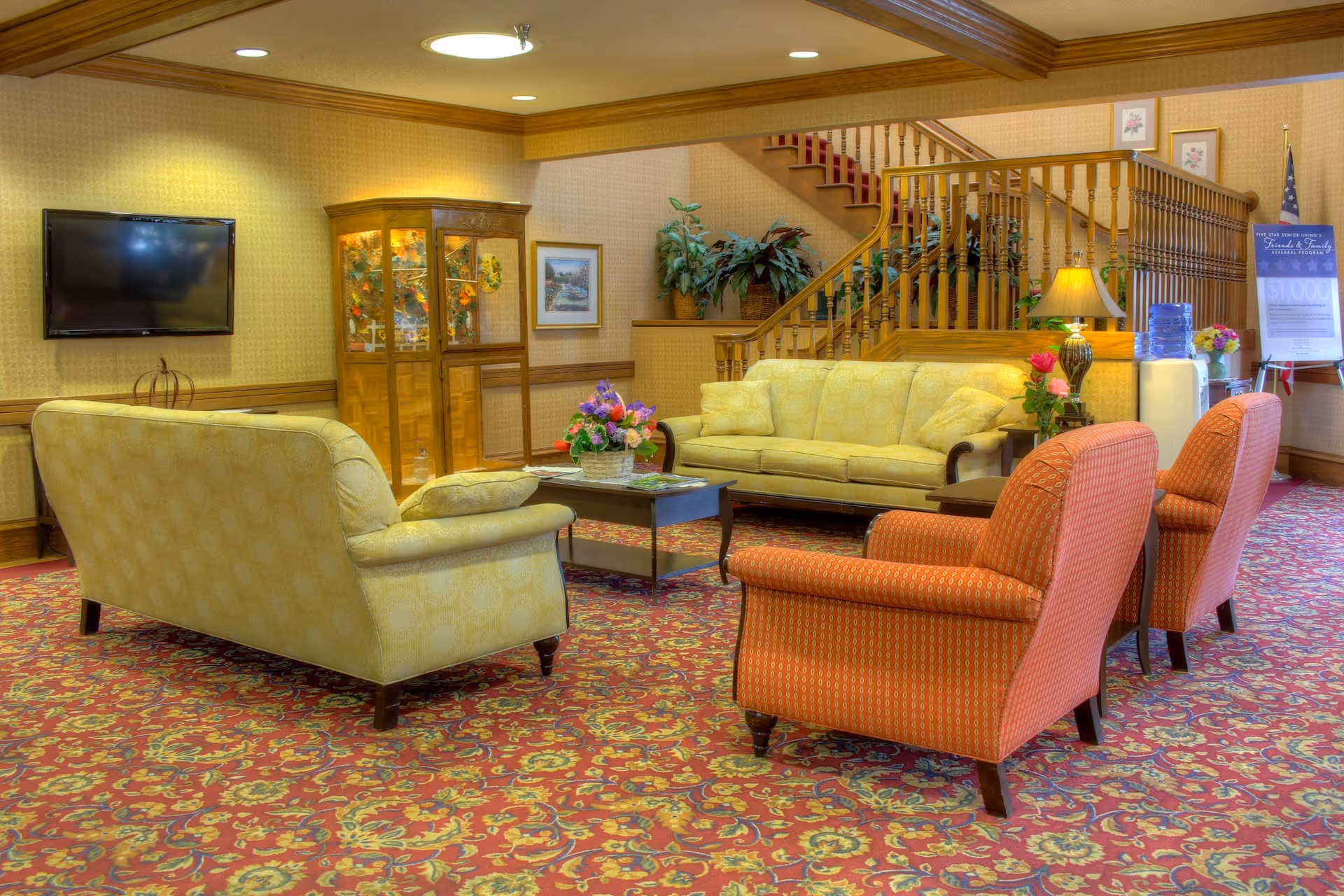 A cozy living room area in an assisted living facility featuring two yellow sofas and two orange armchairs arranged around a dark wooden coffee table with a flower arrangement. The room has a patterned red carpet, a flat-screen TV mounted on the wall, a wooden display cabinet, and a staircase with wooden railings. There are plants and a water dispenser near the staircase, and a signboard is visible in the background.