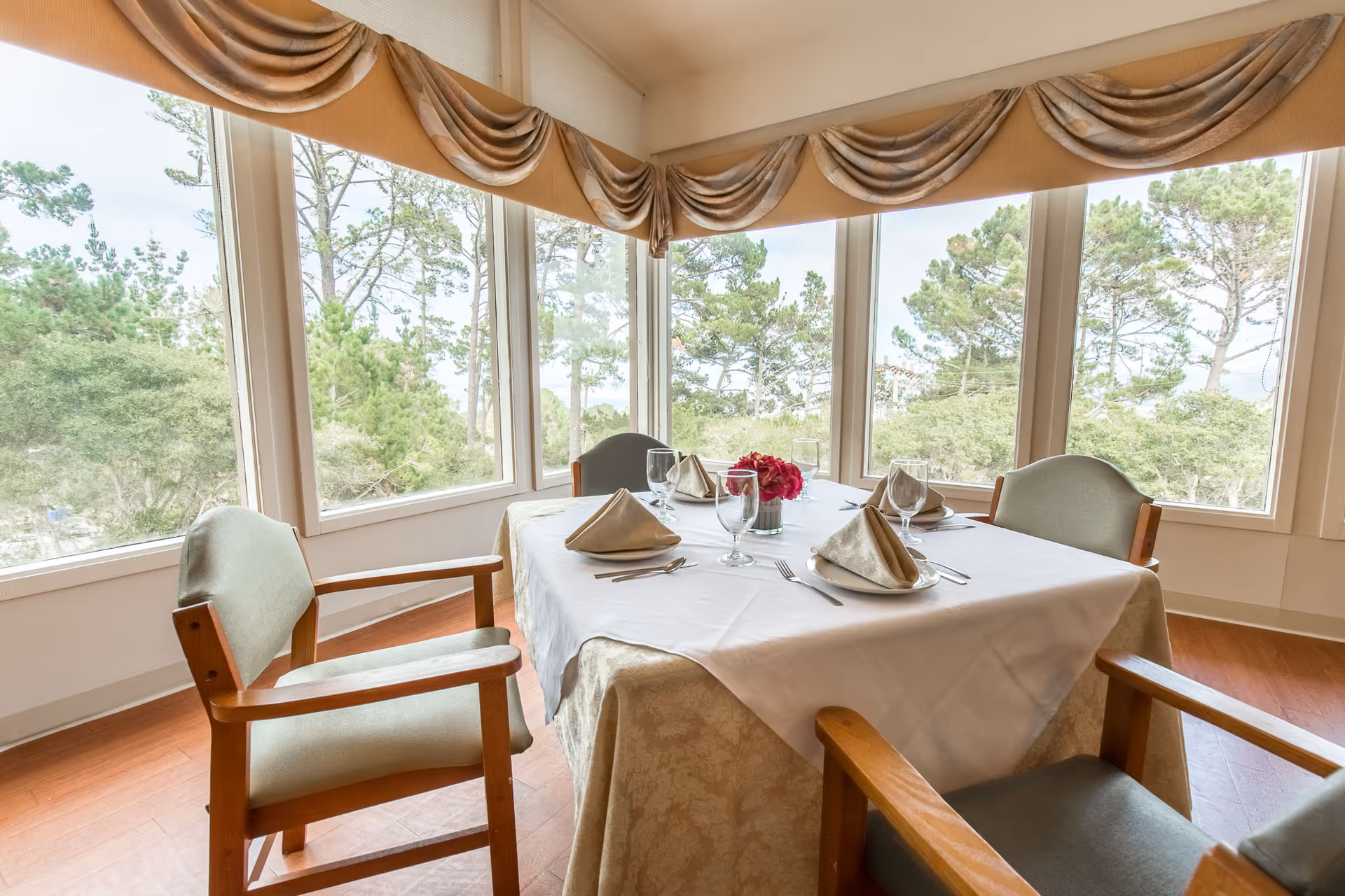A dining table set for four with beige napkins, glassware, and a small flower arrangement in the center, surrounded by wooden chairs with green cushions, positioned next to large windows with a view of trees outside and beige draped valances above.