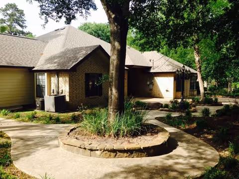 Exterior view of a single-story senior care home building with a light brick facade and multiple roof sections. A large tree is centered in a circular stone planter surrounded by a concrete walkway. The area is landscaped with grass, plants, and trees providing shade.