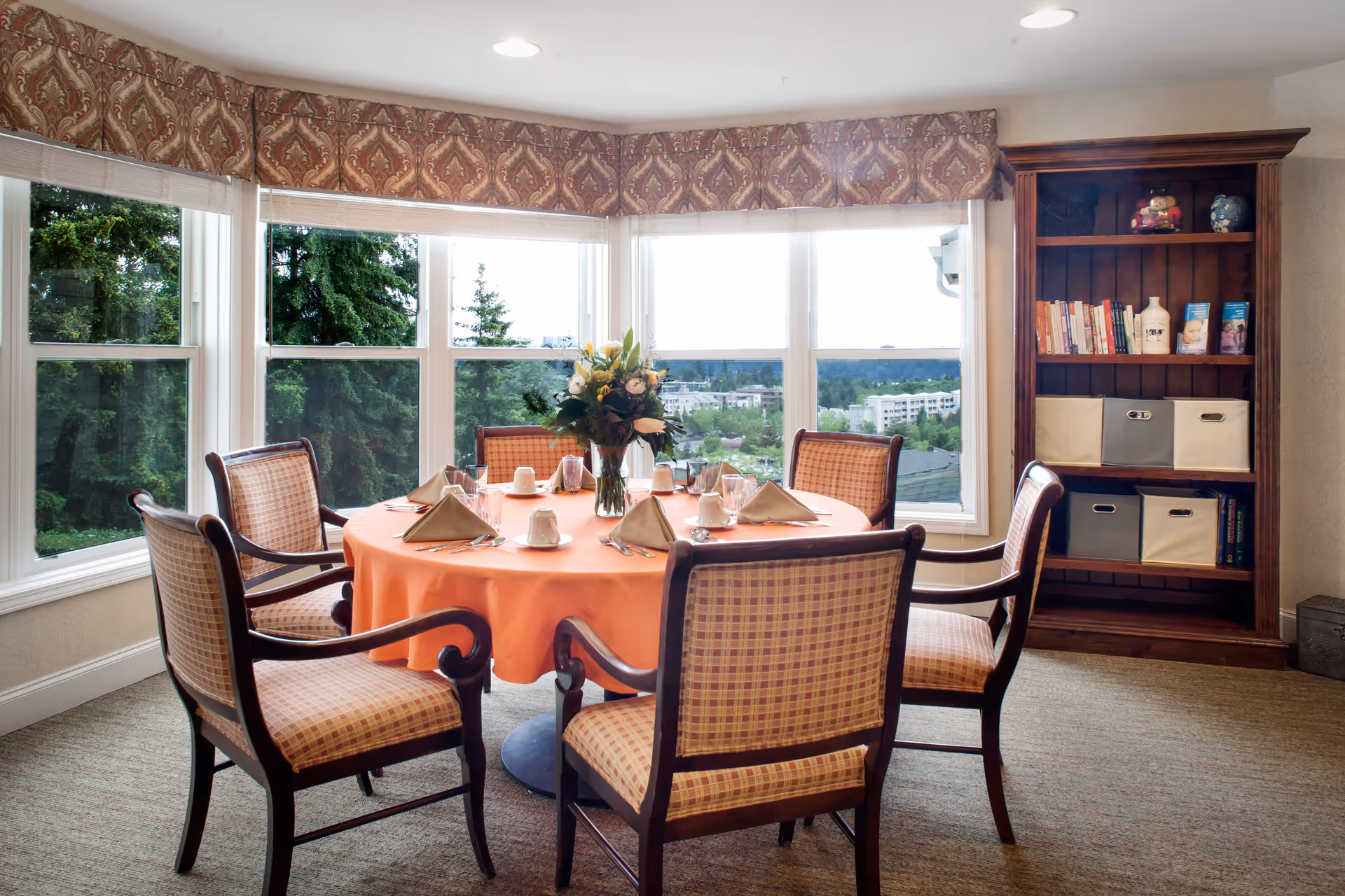 A dining area with a round table covered with an orange tablecloth, set with napkins, cups, glasses, and silverware. Six wooden chairs with plaid upholstery surround the table. A vase with flowers is placed in the center. Large windows behind the table offer a view of trees and buildings outside. To the right, there is a wooden bookshelf with books and storage bins.