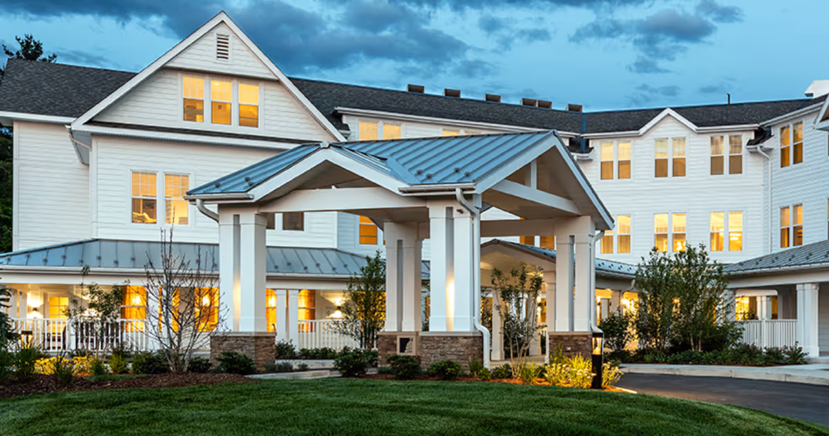 Front exterior of a multi-story white senior living building with a covered porte-cochere and illuminated windows at dusk.