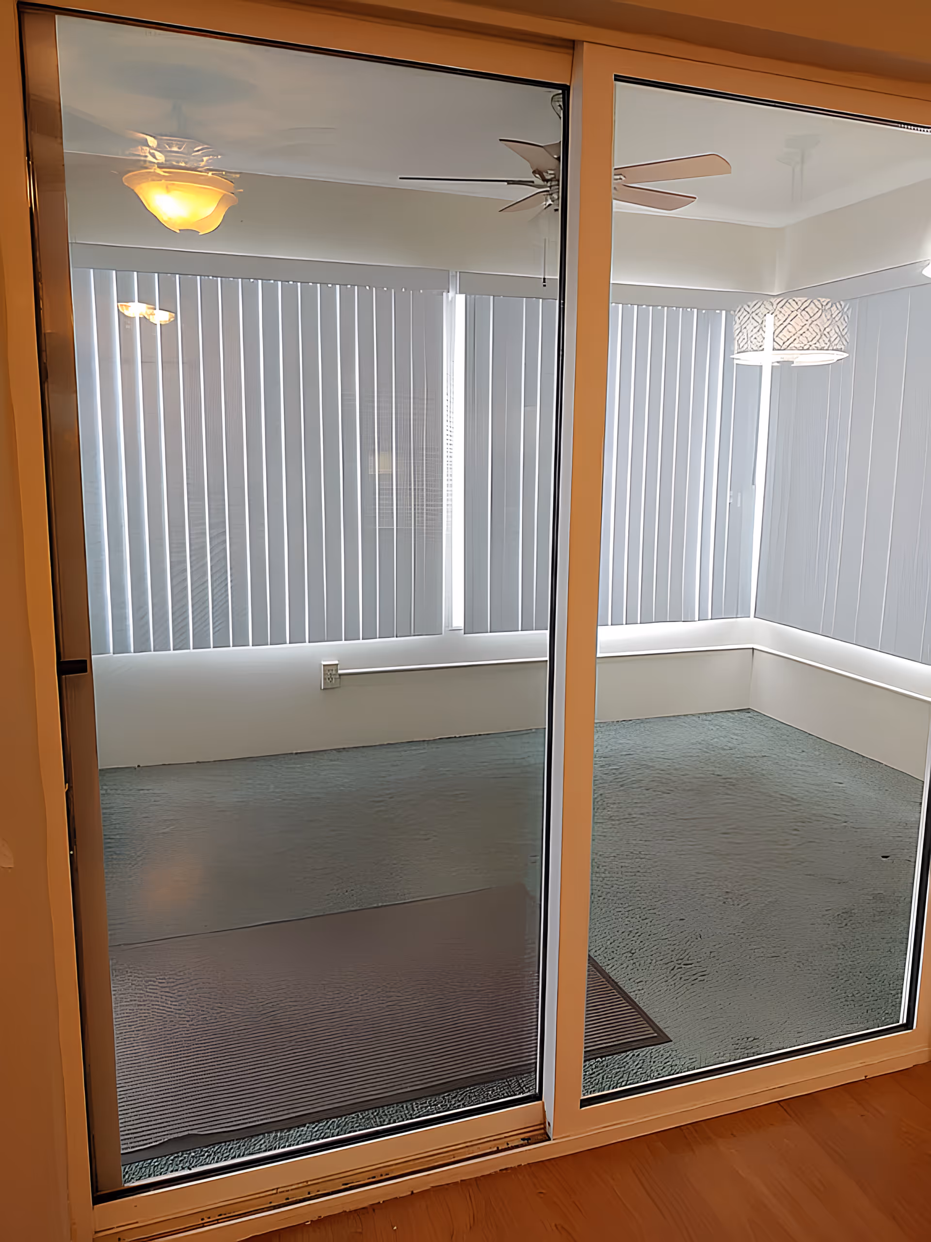 Sliding glass doors open to a bright enclosed sunroom with vertical blinds, ceiling fans, light fixtures, and a carpeted floor.