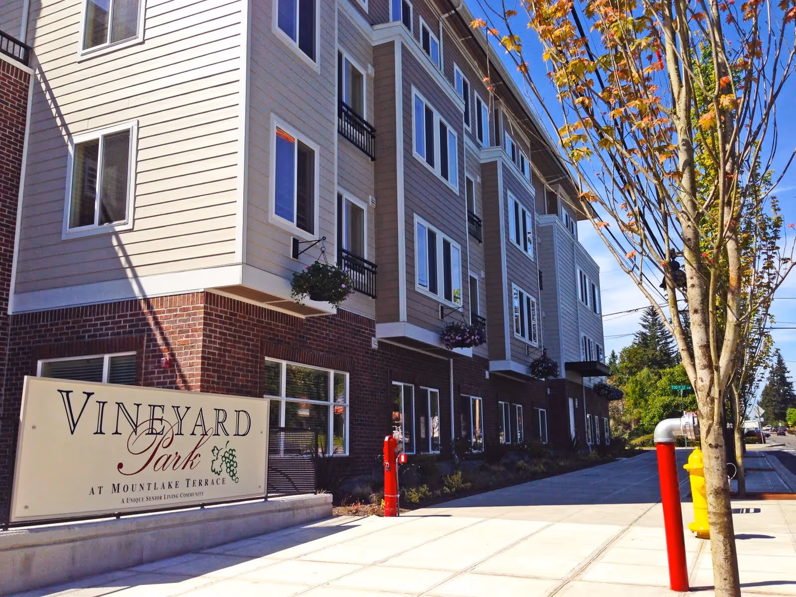 Exterior view of Vineyard Park at Mountlake Terrace, a multi-story senior living community building with beige siding and brick accents. There are several windows, hanging flower baskets, a sidewalk, a tree, and a fire hydrant visible in front of the building under a clear blue sky.