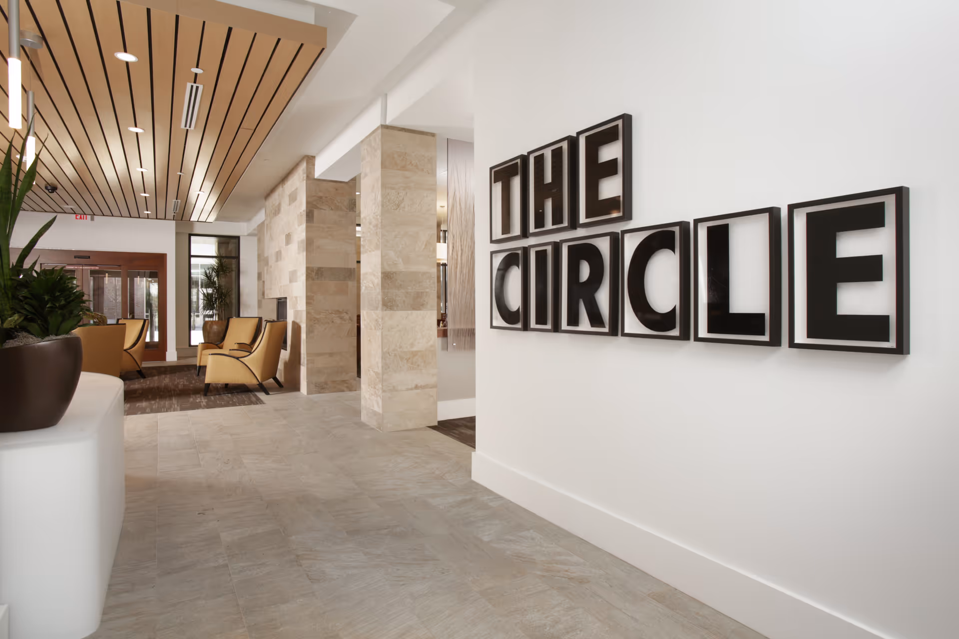 Interior view of a modern senior living facility lobby with beige tiled floor, wooden slatted ceiling, stone pillars, and comfortable tan armchairs. On the right wall, large black letters spell out 'THE CIRCLE'. There are potted plants and large windows near the entrance.