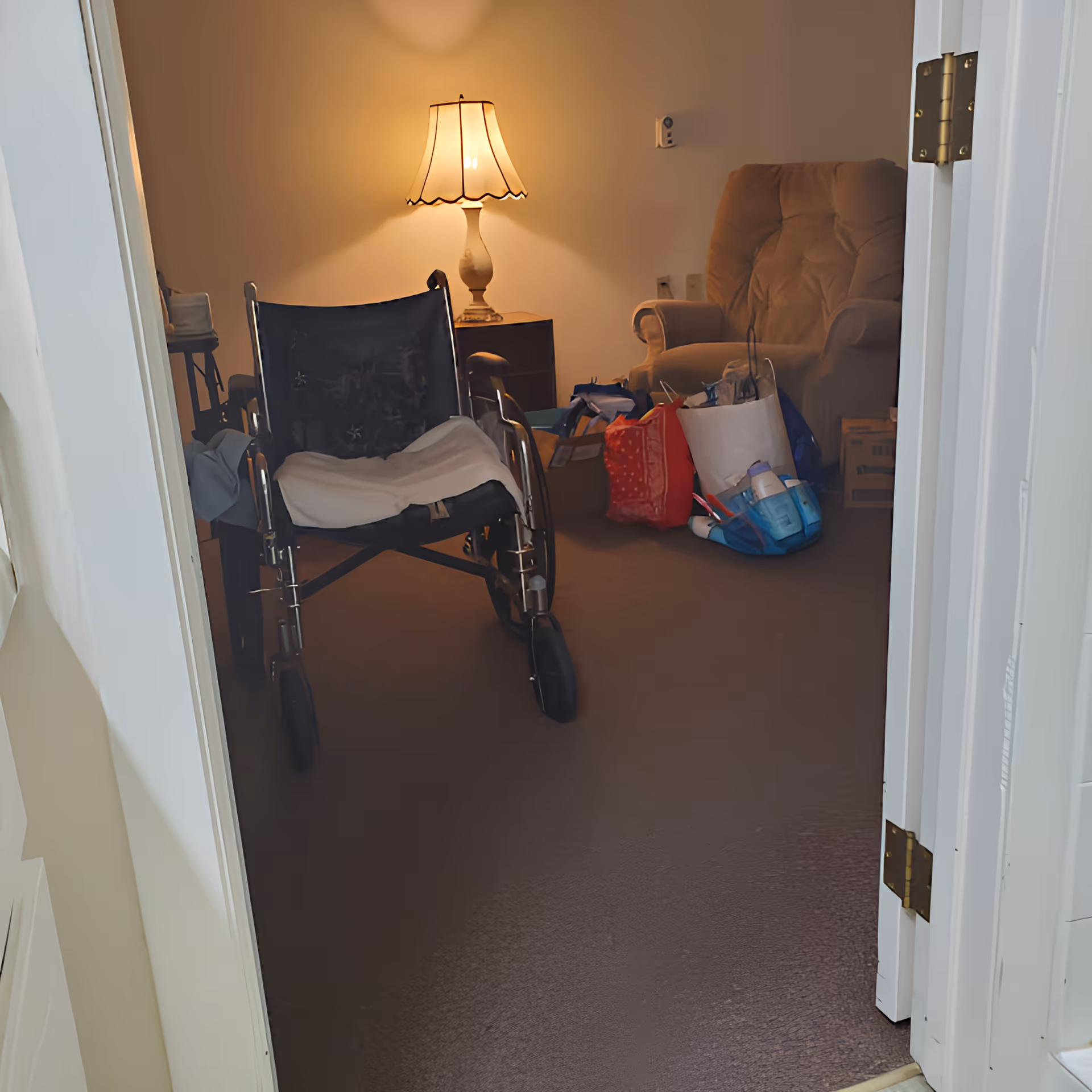 View into a room with a wheelchair in the foreground, a lit table lamp on a small side table, a cushioned armchair, and several bags and boxes on the floor.