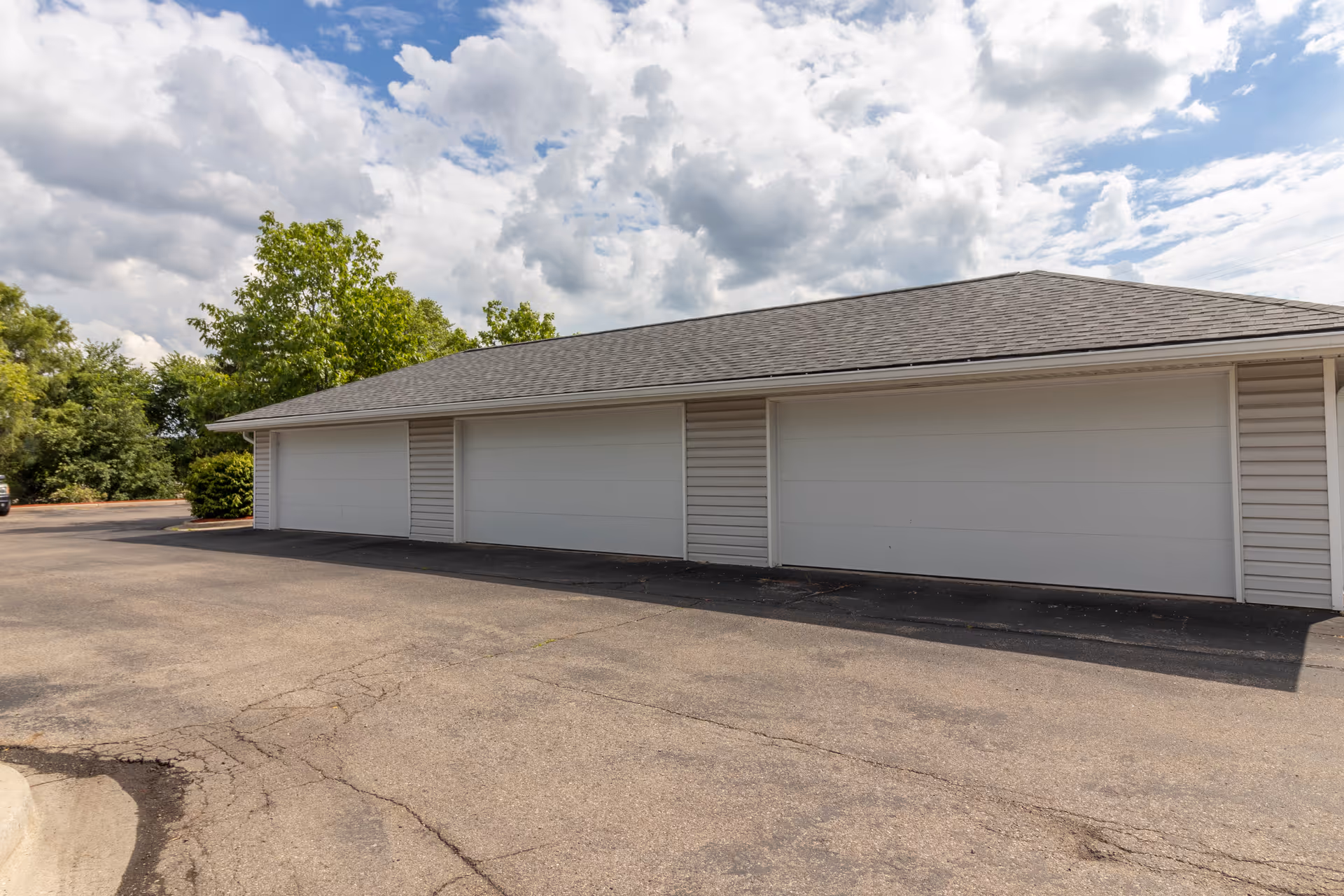 Exterior view of a building with three closed white garage doors, surrounded by trees and under a partly cloudy sky.