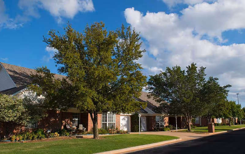 Brick single-story residential building with trees, a manicured lawn, and a sidewalk under a blue sky.