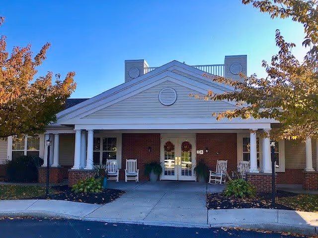 Front exterior view of Celebration Villa of Lebanon PA, showing a building entrance with white columns, a covered porch, rocking chairs, potted plants, and trees with autumn foliage under a clear blue sky.