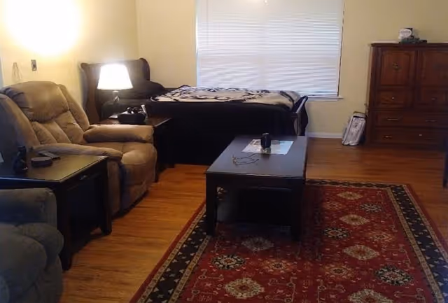 Studio-style living area with two recliners, a coffee table on a patterned rug, and a bed against the back wall beneath a window.