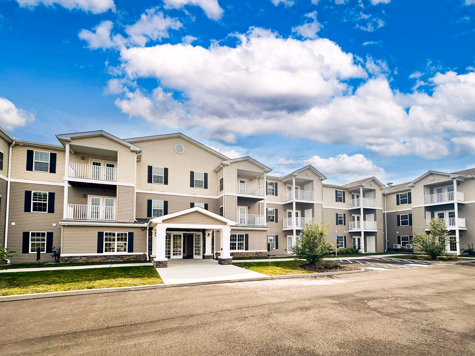 Three-story beige and white senior living building with balconies, a covered entrance, parking area, and blue sky above.