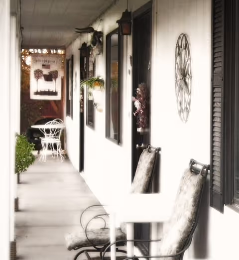 A covered outdoor walkway or porch area with white walls and black window shutters. There are cushioned chairs and a small table along the wall, decorative wall hangings including a clock, and a white metal table with chairs at the far end. Potted plants and hanging planters add greenery to the space.