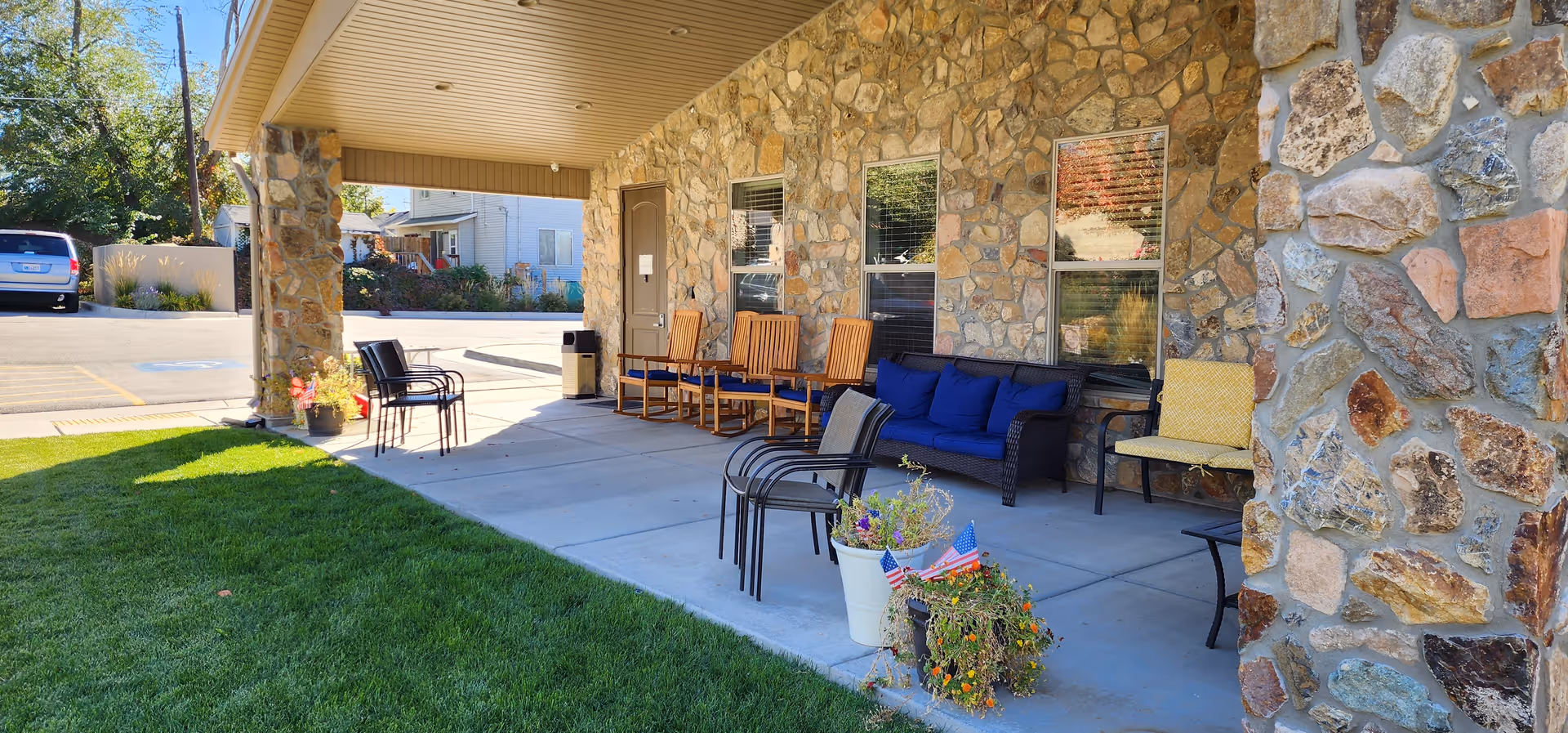 Covered outdoor patio area with stone pillars and walls, featuring several chairs including wooden rocking chairs, cushioned chairs, and a blue cushioned sofa. There are potted plants with flowers and small American flags near the seating area. A parking lot and some houses are visible in the background.