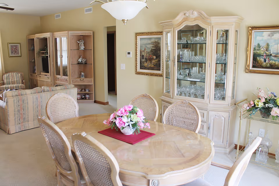 Bright traditional dining room with a round wooden table and floral centerpiece, a glass-front china cabinet, and an adjoining living area.