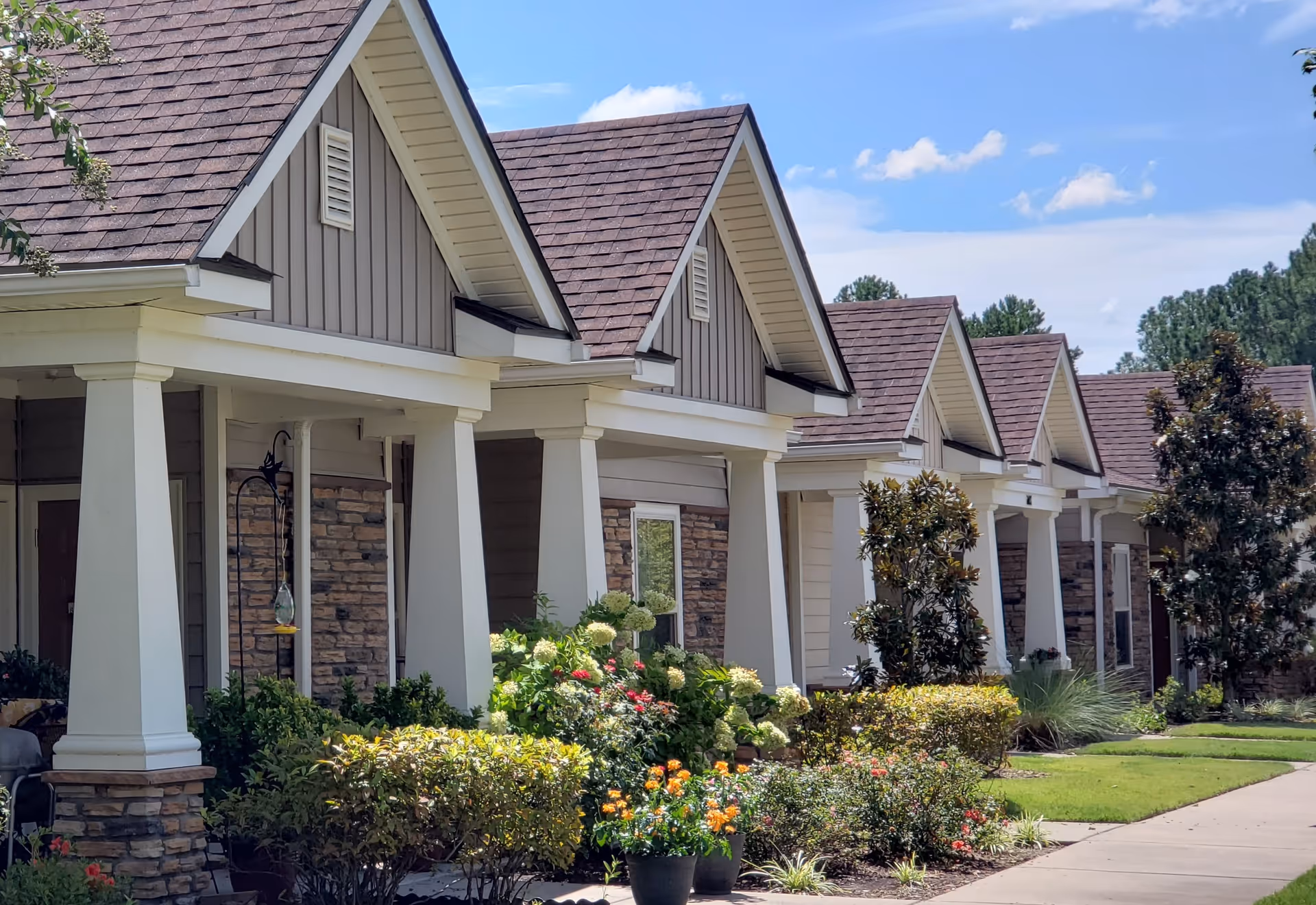 Exterior view of a row of single-story residential buildings with peaked roofs, white columns, and stone accents. The buildings are surrounded by well-maintained landscaping including bushes, flowering plants, and small trees under a blue sky with some clouds.