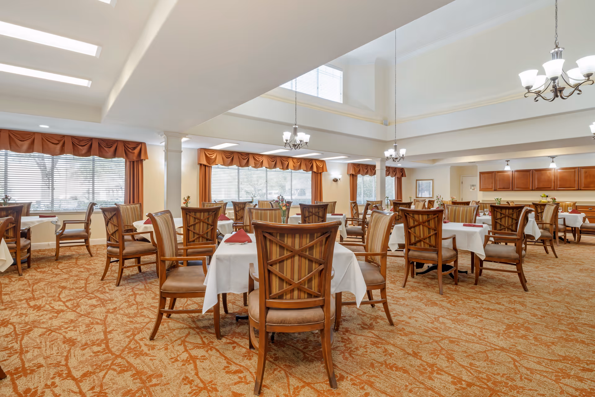 A spacious dining room with multiple tables covered in white tablecloths and set with red napkins. The room features wooden chairs with cushioned seats and striped backs, large windows with orange valances, and warm lighting from chandeliers and ceiling lights. The carpet has an orange and beige pattern, and there are cabinets along one wall.