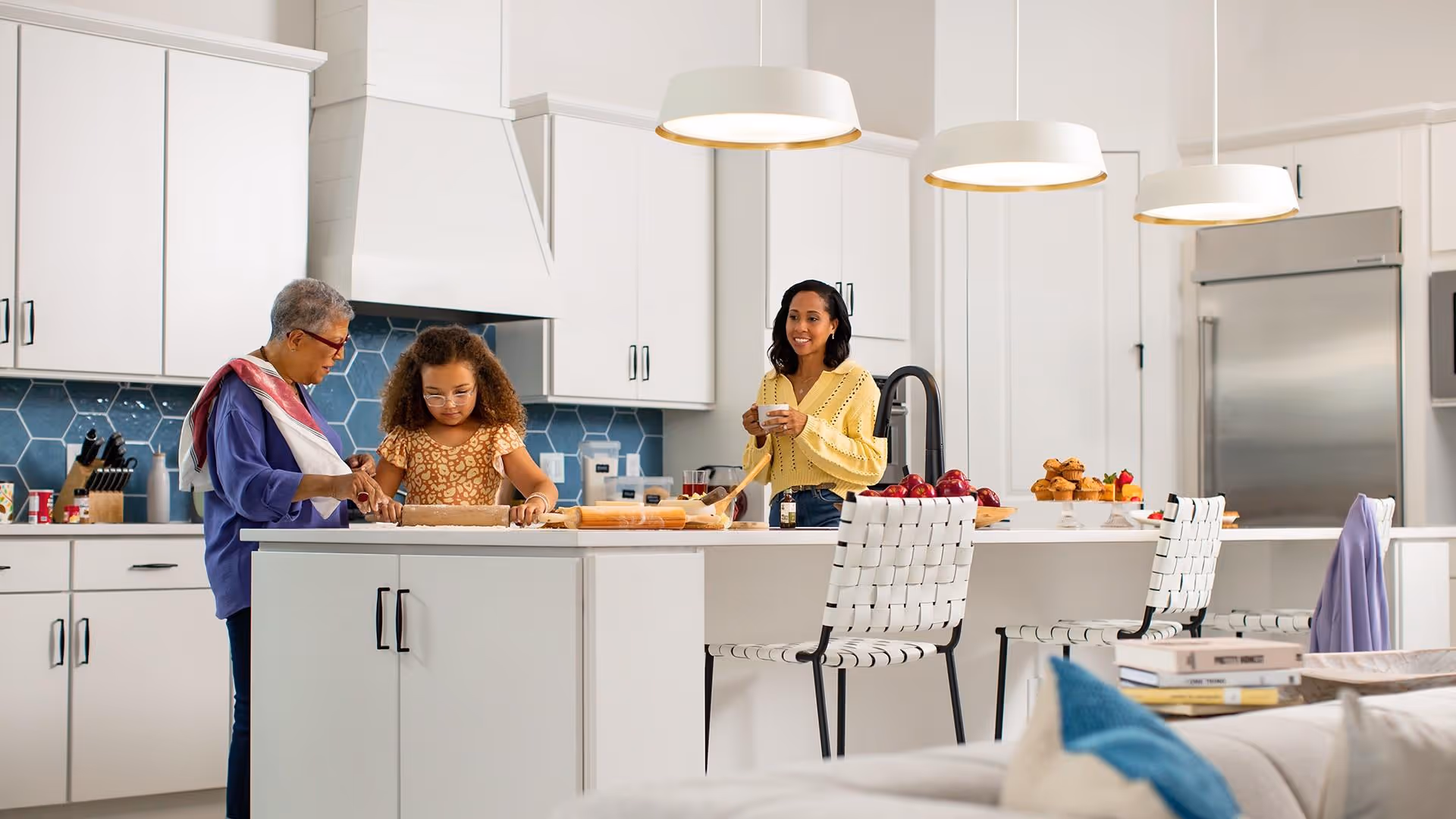 A modern kitchen with white cabinets and a blue hexagonal tile backsplash. An older woman and a young girl are rolling dough on the kitchen island, while a younger woman stands nearby holding a cup. The kitchen island has white woven chairs and is decorated with apples and muffins. Pendant lights hang above the island.