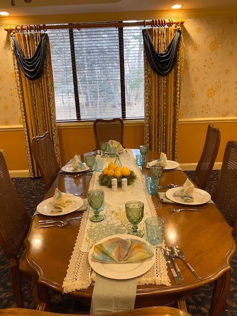 A formal dining room with a wooden table set for six people. The table is decorated with a lace-edged table runner, green glassware, folded napkins on white plates, and a centerpiece of lemons and greenery. The room has floral wallpaper, yellow walls with white trim, and a large window with patterned curtains and dark valances.