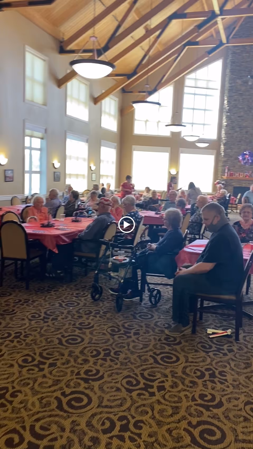 Sunlit common dining room with seniors seated at round tables beneath a high vaulted wood ceiling.