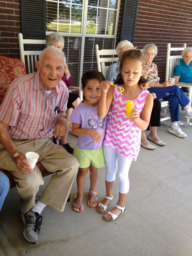 An elderly man sitting on a bench outside a brick building with several elderly women seated in rocking chairs behind him. Two young girls stand in front of the man, one blowing bubbles while the other smiles at the camera.