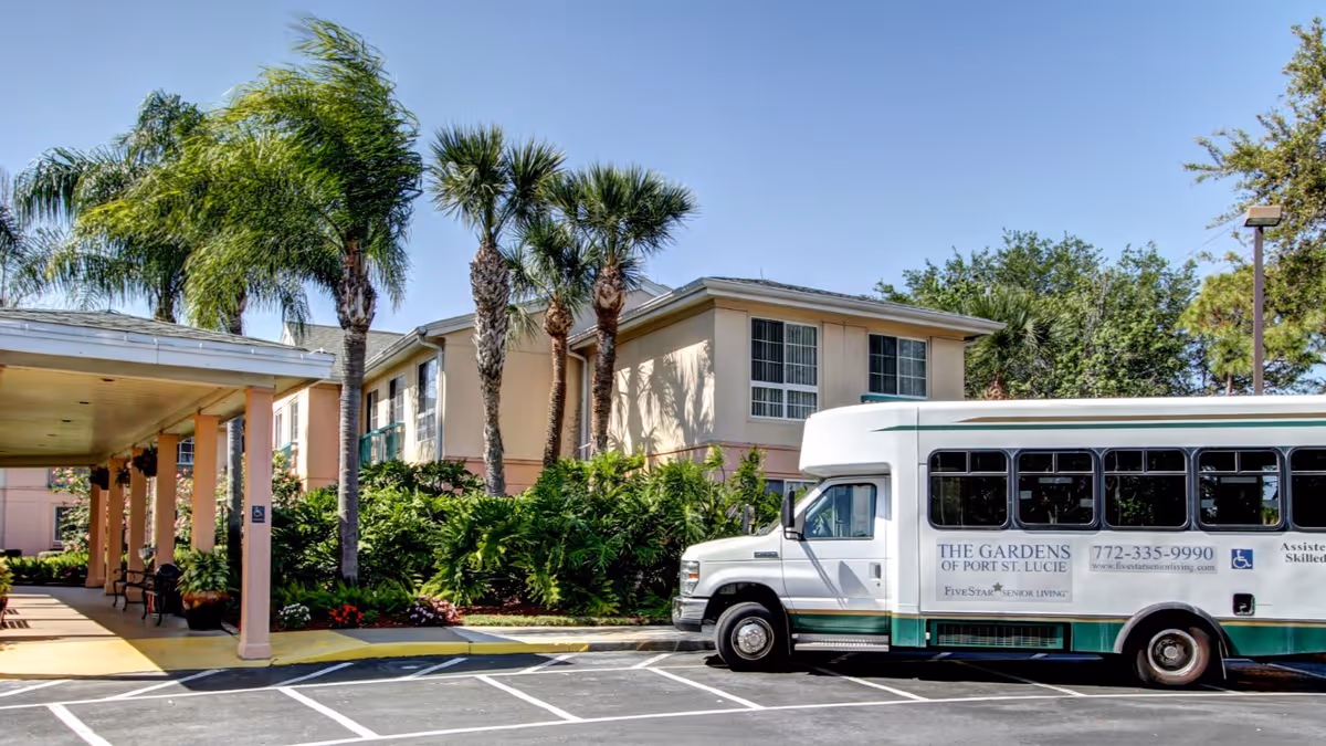Entrance of The Gardens of Port St. Lucie senior living building with a shuttle bus parked in front and palm trees lining the driveway.
