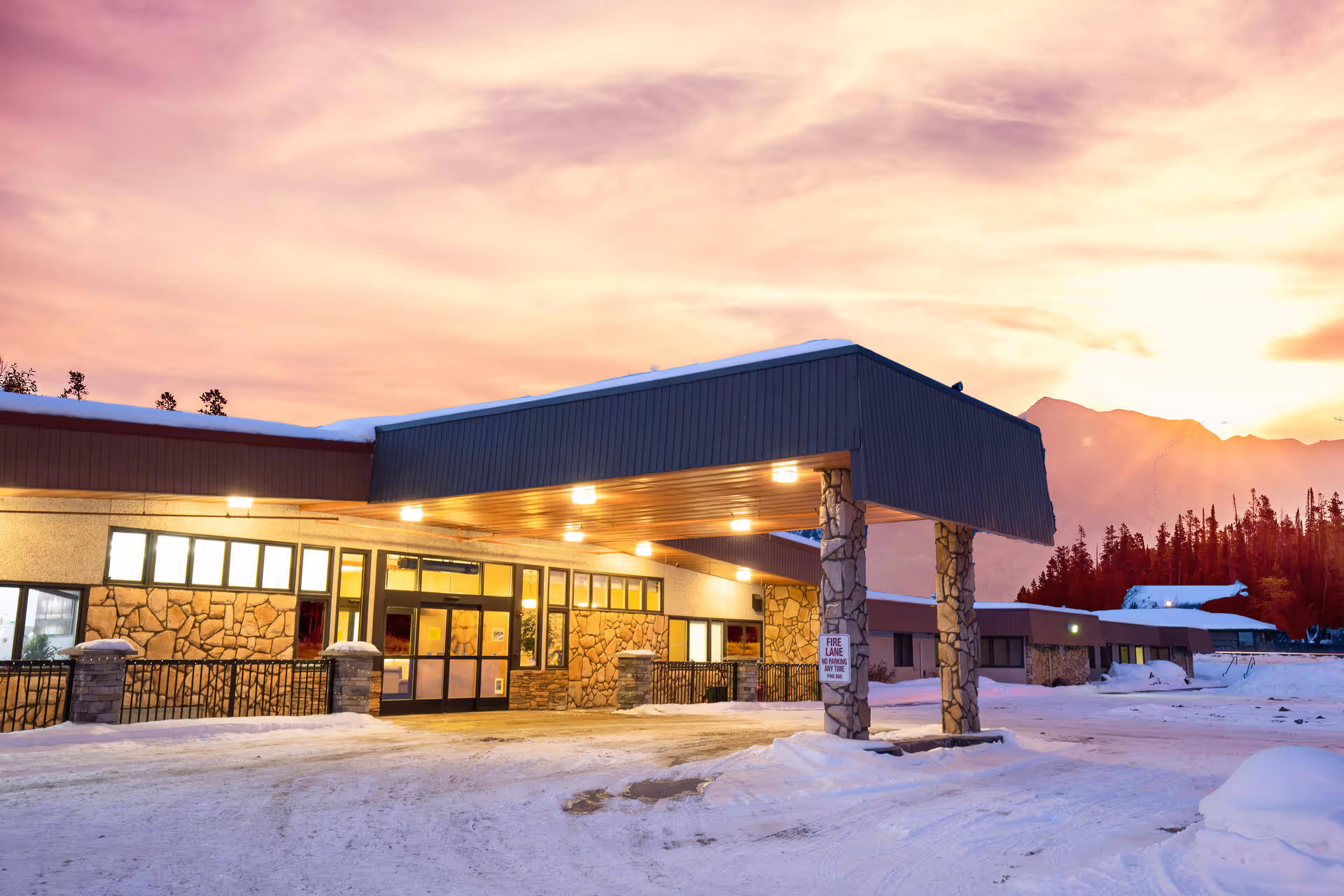 Snow-covered driveway and illuminated covered entrance of a single-story health facility at sunset with mountains in the background.