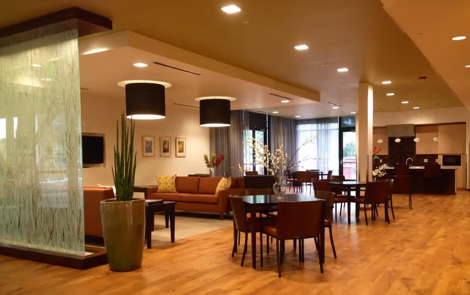 A spacious and well-lit common area in a senior living facility featuring wooden floors, round tables with chairs, a seating area with brown sofas and yellow pillows, decorative plants, modern hanging light fixtures, and large windows with curtains allowing natural light.