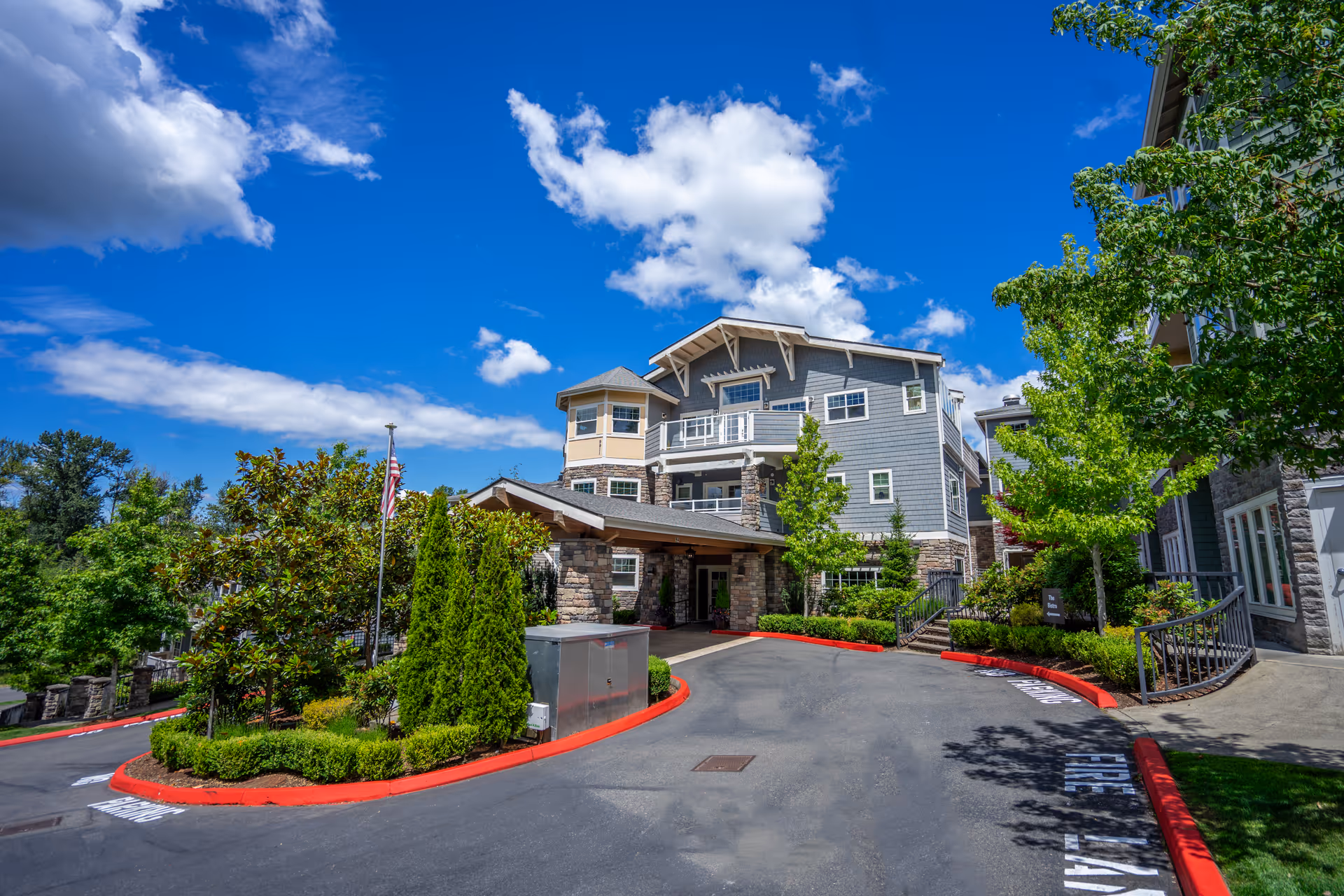 Exterior view of Chateau Bothell Landing Retirement facility on a sunny day with a clear blue sky and some clouds. The building is multi-story with gray siding, stone accents, and balconies. There is a driveway with red curbs, landscaped greenery including trees and bushes, and an American flag near the entrance.