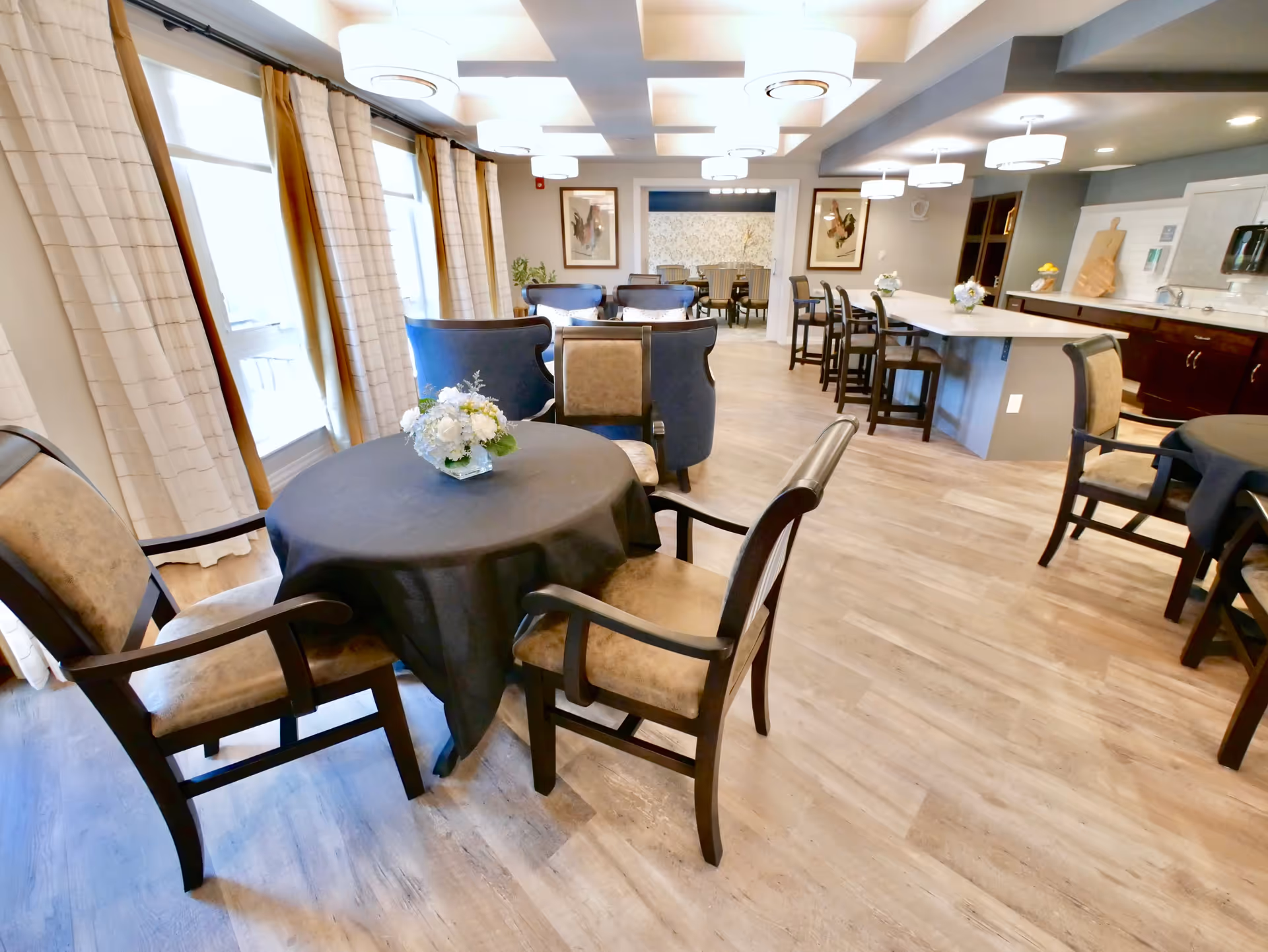 Bright communal dining and lounge area with round tables, upholstered chairs, a kitchen island, and pendant lighting.