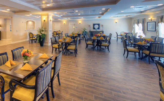 A spacious and well-lit dining room in a senior living facility with multiple tables set with yellow napkins and floral centerpieces. The room features wood-look tile flooring, beige walls, and large windows with curtains allowing natural light to enter. Comfortable chairs surround each table, and decorative plants and wall sconces add to the inviting atmosphere.
