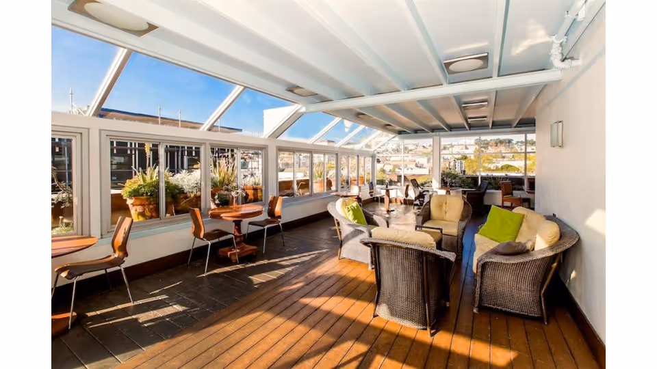 Sunlit enclosed rooftop lounge with wicker sofas, chairs, and small tables overlooking the city.