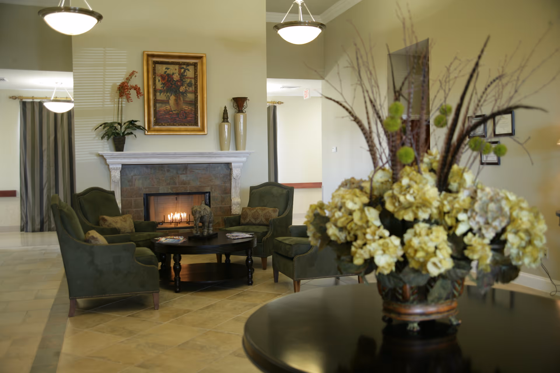 A cozy living room area with four green upholstered armchairs arranged around a round wooden coffee table in front of a lit fireplace. Above the fireplace is a framed painting of flowers. There are decorative vases on the mantel and a large floral arrangement on a round table in the foreground. The room has beige walls and tiled flooring, with soft lighting from ceiling fixtures.