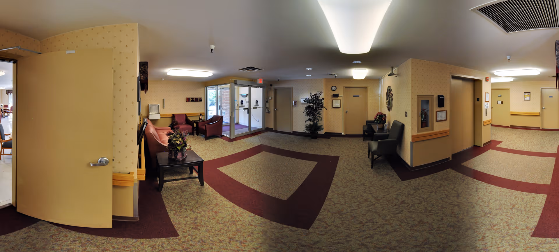 A spacious senior living facility common area with patterned carpet and beige walls. The room features several seating areas with chairs and a sofa, a coffee table with a flower arrangement, potted plants, and wall decorations. There are double glass doors leading outside and an elevator on the right side. The area is well-lit with ceiling lights.