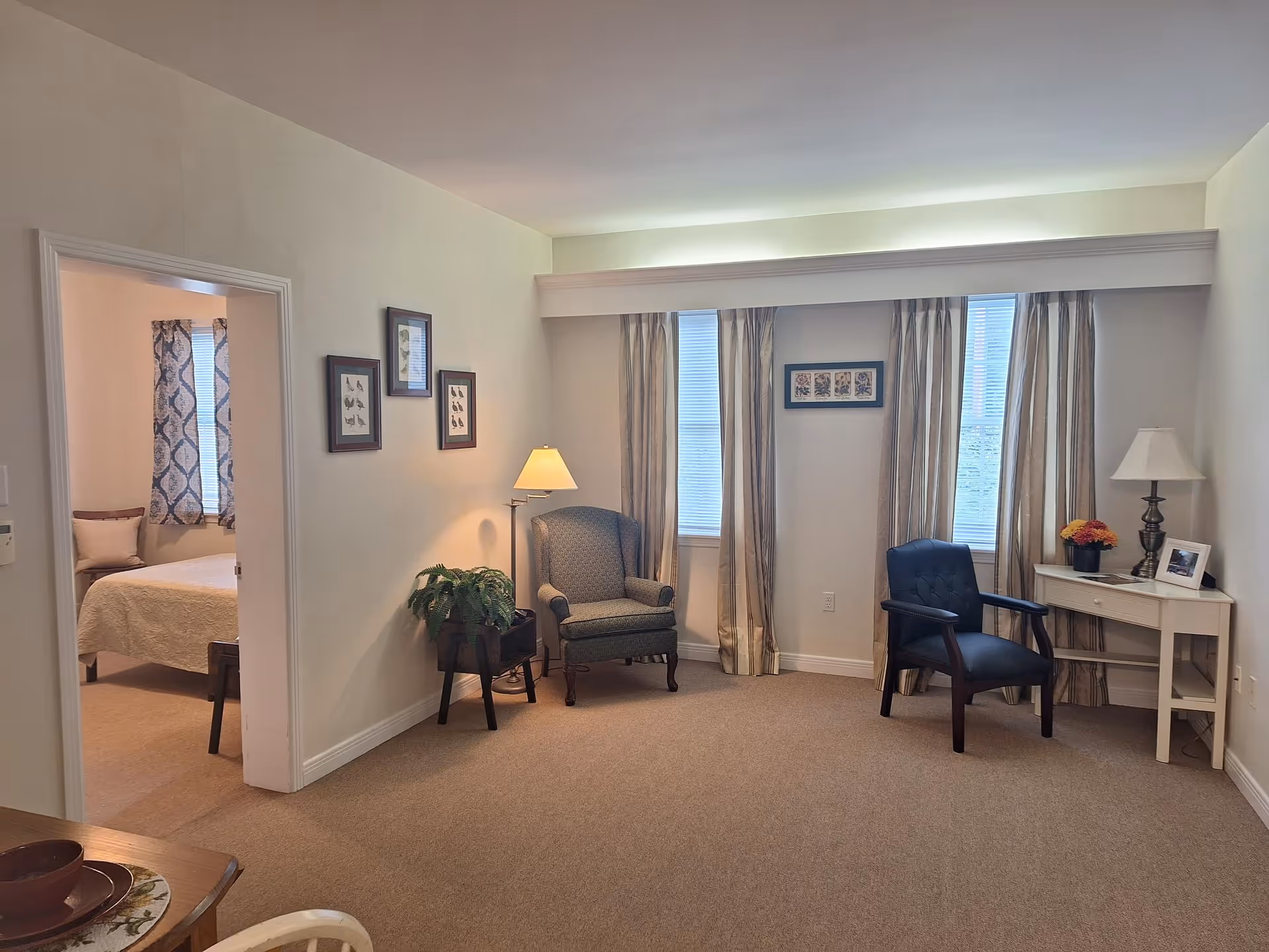 A cozy living room area with two armchairs, a small white desk with a lamp and a flower pot, and three windows with beige curtains. The walls are light-colored and decorated with framed pictures. To the left, there is an open doorway leading to a bedroom with a bed and patterned curtains.