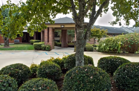Brick building entrance with a covered walkway, trees, and manicured shrubs.