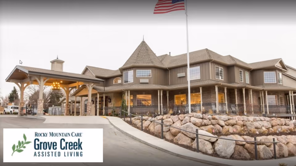 Front exterior of a large two-story assisted living building with a covered entrance, American flag, rock landscaping, and a Grove Creek sign.