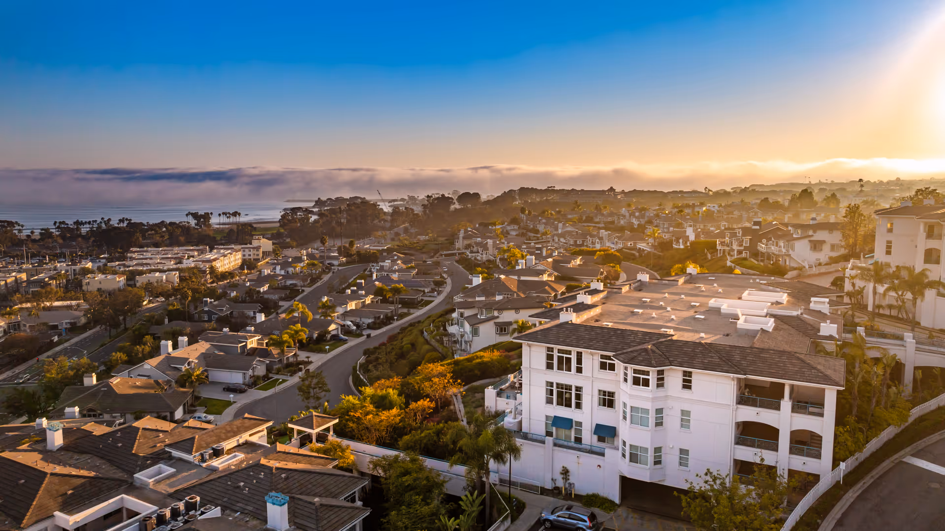 Aerial view of a coastal residential neighborhood at sunset with multiple houses, trees, and a large white building in the foreground. The ocean and a layer of fog are visible in the background under a clear blue sky.