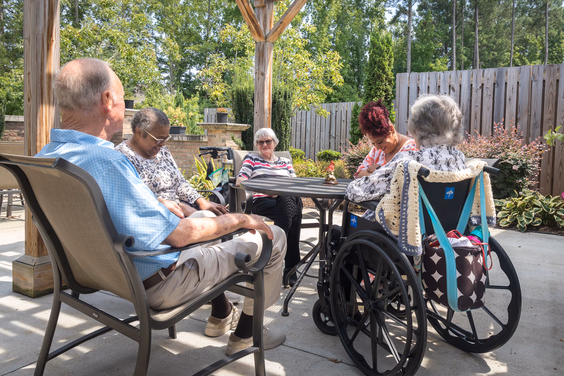 A group of elderly people sitting and socializing outdoors under a wooden pergola. Two women are in wheelchairs, and the others are seated in patio chairs around a round metal table. The setting is a garden area with plants, trees, and a wooden fence in the background.